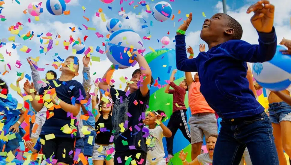 Children jump and play with beach balls as colorful confetti fills the air at an outdoor inflatable festival.