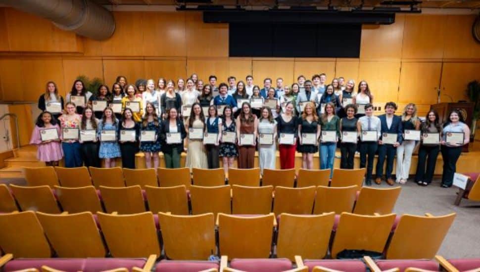Large group of West Chester University students standing on a stage in an auditorium, each holding a certificate, during the 2026 1871 Award ceremony, with rows of empty theater seats visible in the foreground and a wood-paneled backdrop behind them.