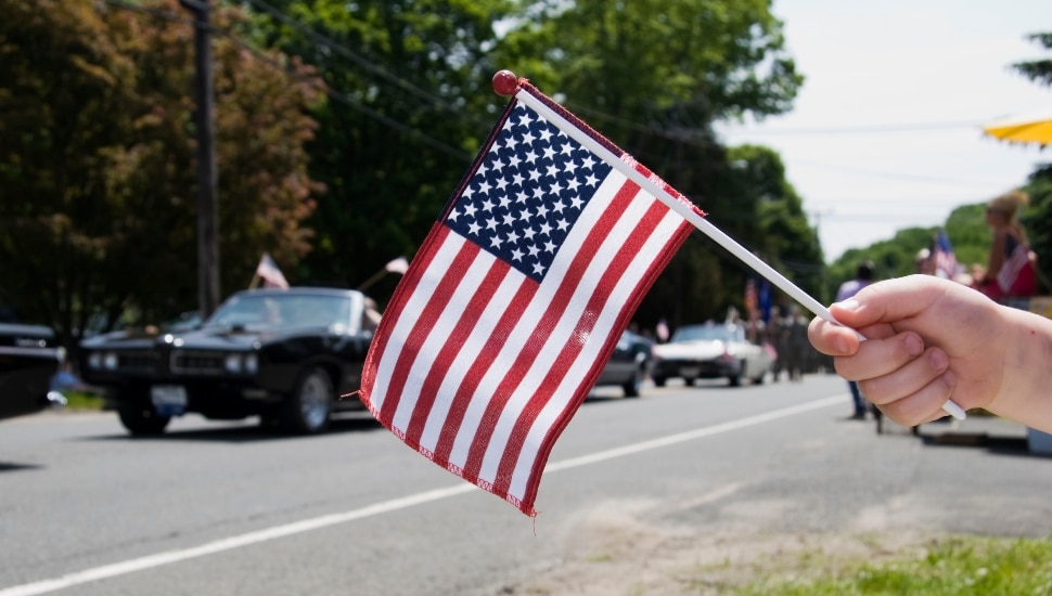 A hand waves a miniature American flag in front of a Memorial Day parade.