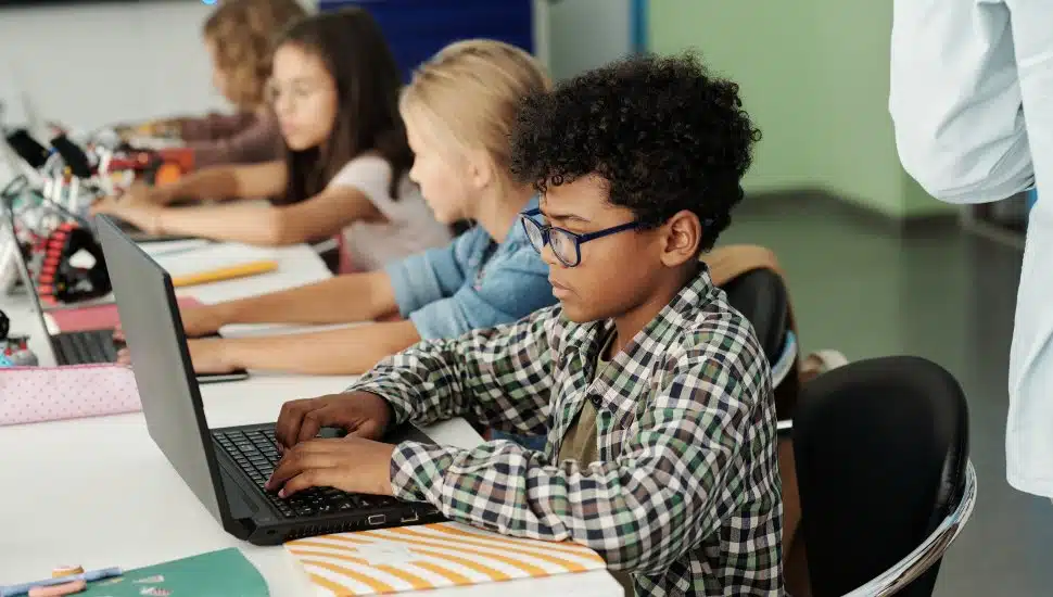 Side view of youthful schoolboy and his classmates working in front of laptops.