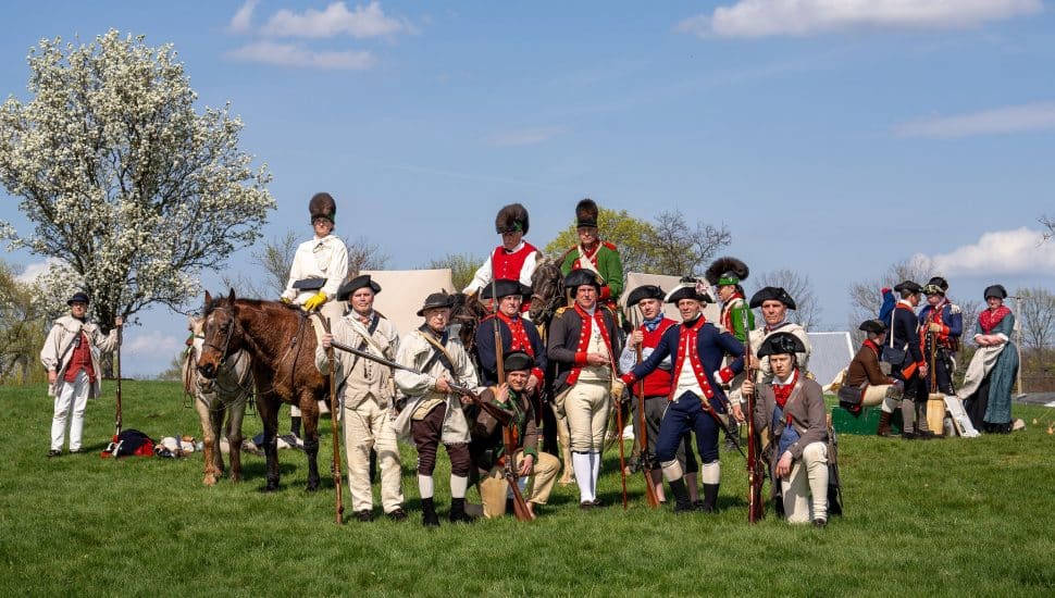 Group of Revolutionary War reenactors in period uniforms with muskets and two horses on a grassy field at Pottsgrove Manor in Pottstown.