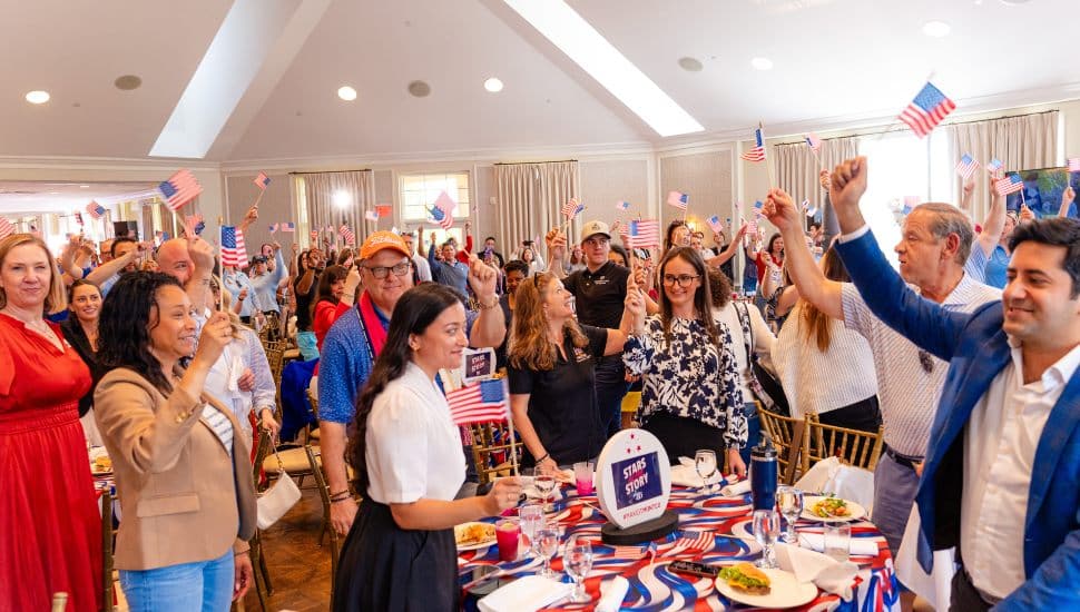 A crowded ballroom of smiling attendees wave small American flags at the VFTCB 2026 Travel Rally "Stars of the Story" event at Whitemarsh Valley Country Club in Montgomery County.