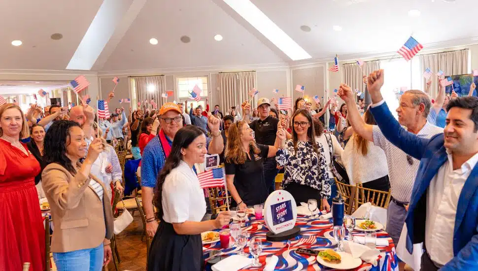 A crowded ballroom of smiling attendees wave small American flags at the VFTCB 2026 Travel Rally "Stars of the Story" event at Whitemarsh Valley Country Club in Montgomery County.