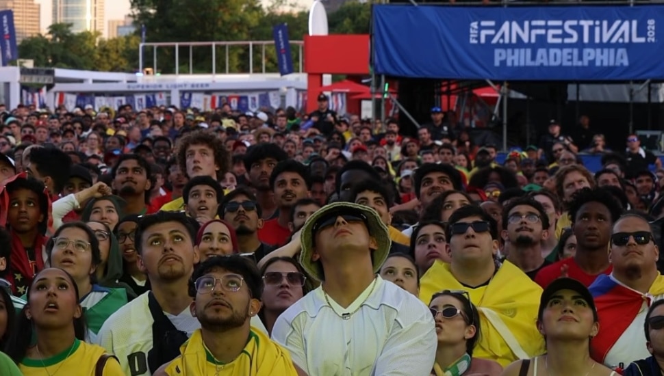 A dense crowd of soccer fans, many wearing team colors and sunglasses, looks up at a big screen at the FIFA Fan Festival in Philadelphia, with the event's blue banner visible in the background.
