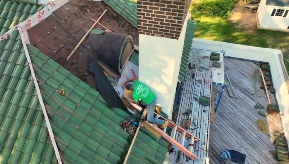 An O'Donnell Roofing crew member works on a green tile roof replacement near a brick chimney, viewed from above.