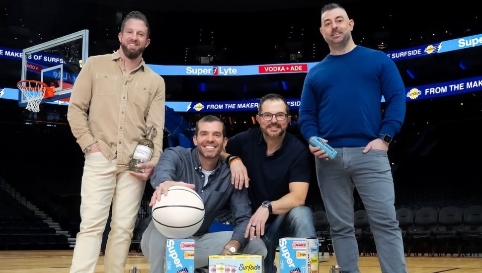 The four founders of Stateside Spirits pose on a basketball court holding a Stateside Vodka bottle and Super Lyte cans, with Surfside and Super Lyte product boxes arranged in front of them and arena LED displays promoting the Super Lyte Vodka + ADE launch in the background.