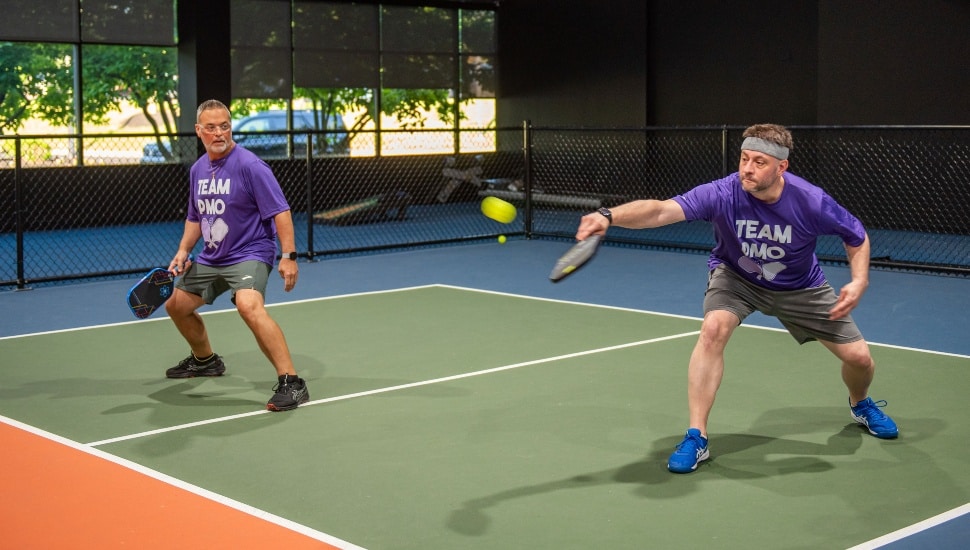 Two middle-aged men in purple shirts play pickleball.