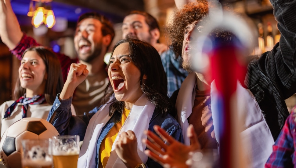 Excited friends cheering for their football team at a bar counter with beer glasses and a soccer ball, celebrating a goal victory.