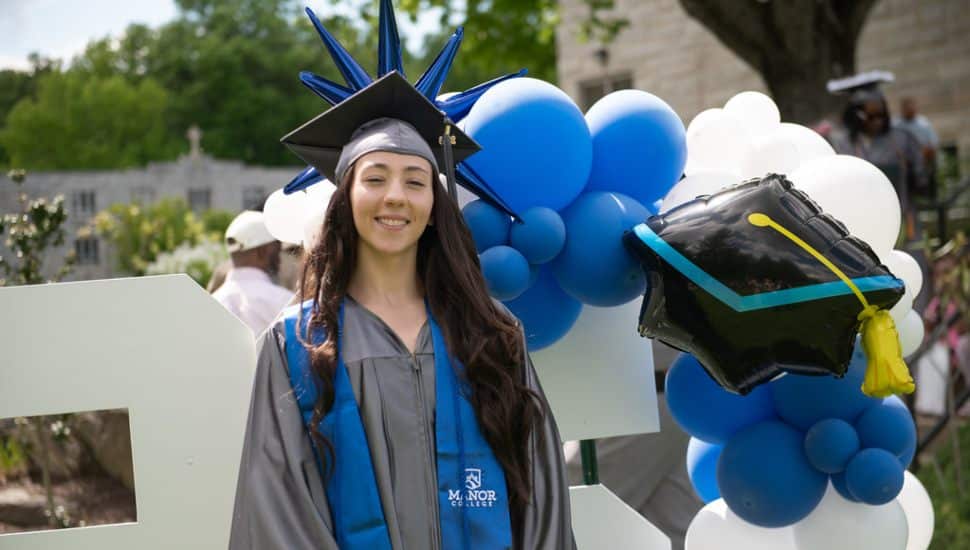 Manor College graduate in cap and gown smiling outdoors at the 75th Commencement ceremony.